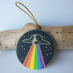 May include: A round, black and white ornament with a gold string. The ornament features a black and white illustration of a UFO with a rainbow beam of light coming from it.
