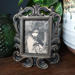 May include: A black ornate picture frame with a vintage black and white photo of a person wearing a witch's hat and holding a cat.