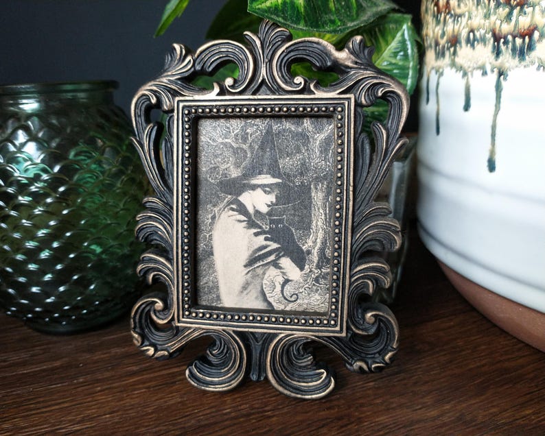 May include: A black ornate picture frame with a vintage black and white photo of a person wearing a witch's hat and holding a cat.