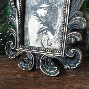 May include: A black ornate picture frame with a gold-toned finish. The frame holds a black and white photograph of a person with a hat and a coat.