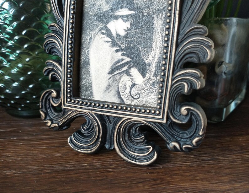 May include: A black ornate picture frame with a gold-toned finish. The frame holds a black and white photograph of a person with a hat and a coat.