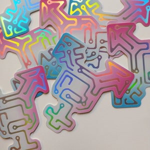 Many identical stickers in the shape of a transgender symbol, with the symbol made of a circuit board pattern. The circuits are holographic rainbow, and the surrounding area is a soft gradient of blue to white to pink, the colors of the trans flag.