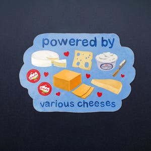 Può includere: Un adesivo azzurro con il testo "powered by various cheeses" in blu scuro. Il design presenta illustrazioni di una forma di brie, formaggio svizzero, una vaschetta di formaggio spalmabile con coltellino, due Gouda rossi e blocchi di cheddar.