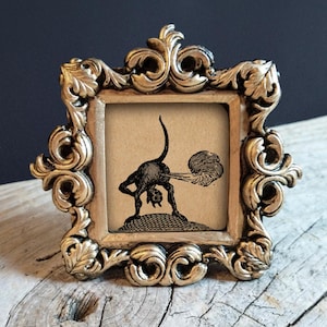 Puede incluir: Un pequeño marco dorado ornamentado con una ilustración vintage en blanco y negro de un gato. El gato está doblado, con una forma de nube que emana de su parte trasera. El marco tiene un diseño detallado de volutas.