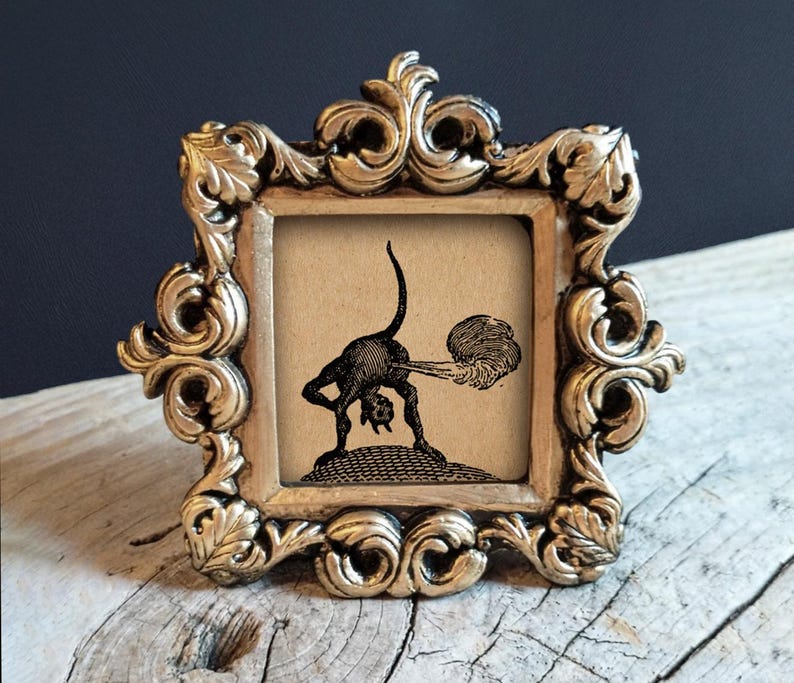 May include: A small square frame with an ornate gold-colored metallic border featuring intricate scrollwork and leaf motifs. Inside the frame is a black and white illustration of a small horned creature bending over with a cloud of smoke behind it.