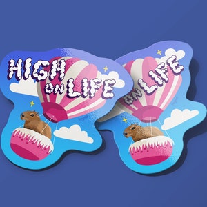 May include: Two stickers featuring a cartoon capybara riding in a hot air balloon. The balloon is pink and white with the words "High on Life" in a bubble font. The stickers are on a blue background.