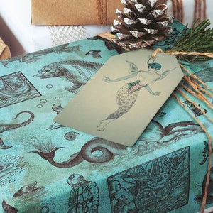May include: Gift-wrapped present with teal wrapping paper featuring illustrations of sea creatures and ships. A mermaid tag is attached with twine. A pine cone and greenery add a festive touch.