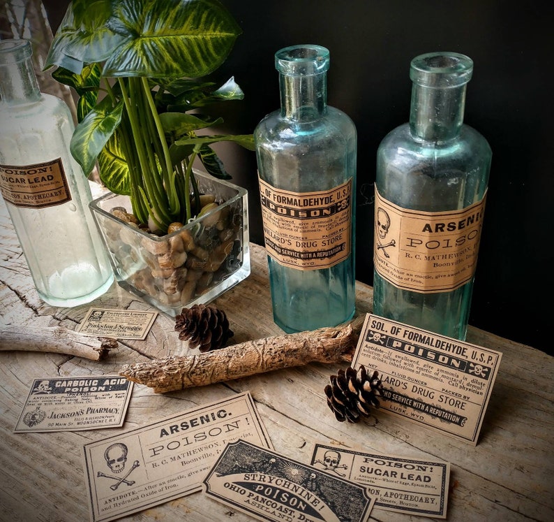 A scene showing antique reproduction label stickers from the Poison Bottle Labels sticker pack. The stickers are printed with blank ink on light brown kraft paper and are rectangular. Formaldehyde and arsenic stickers are shown on antique blue glass pharmacy bottles. Additional stickers such as sugar lead and strychnine are shown lying on a rustic wood surface.