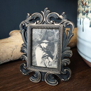 May include: A black ornate picture frame with a gold-toned finish holds a black and white image of a person wearing a hat and cloak. The image is set against a dark background.