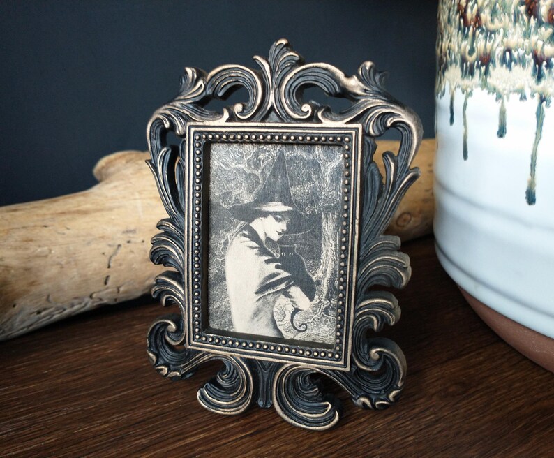 May include: A black ornate picture frame with a gold-toned finish holds a black and white image of a person wearing a hat and cloak. The image is set against a dark background.