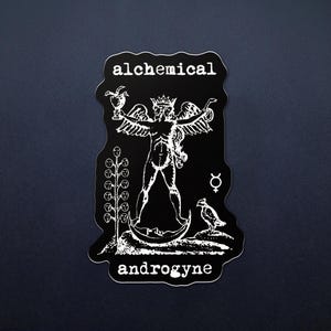 May include: Black sticker featuring a white illustration of a winged figure with a chalice and a bird. The words "alchemical" and "androgyne" are printed in white. The sticker has an irregular, distinctive shape.