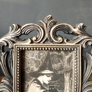 May include: A black and gold ornate picture frame with a black and white image of a woman in a witch's hat holding a black cat.