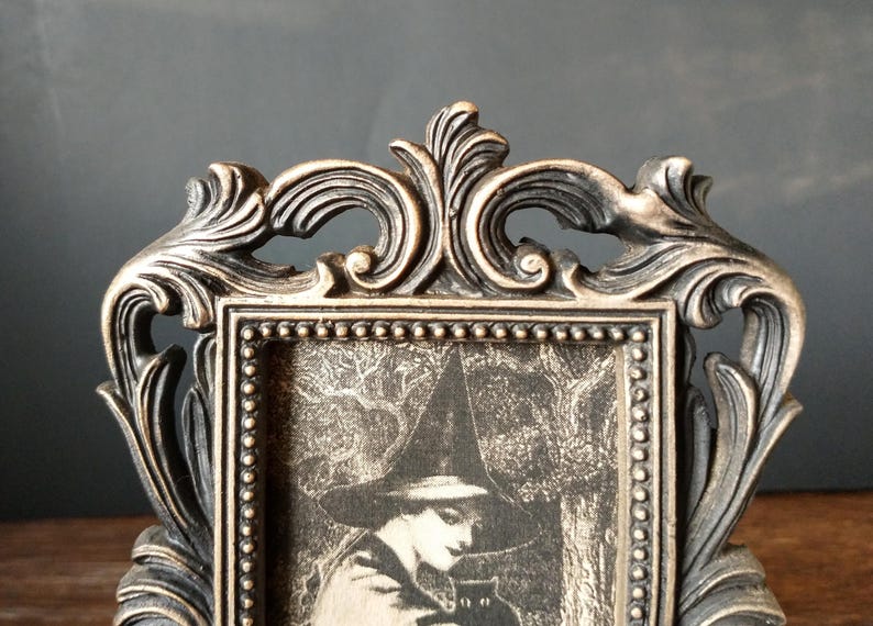 May include: A black and gold ornate picture frame with a black and white image of a woman in a witch's hat holding a black cat.
