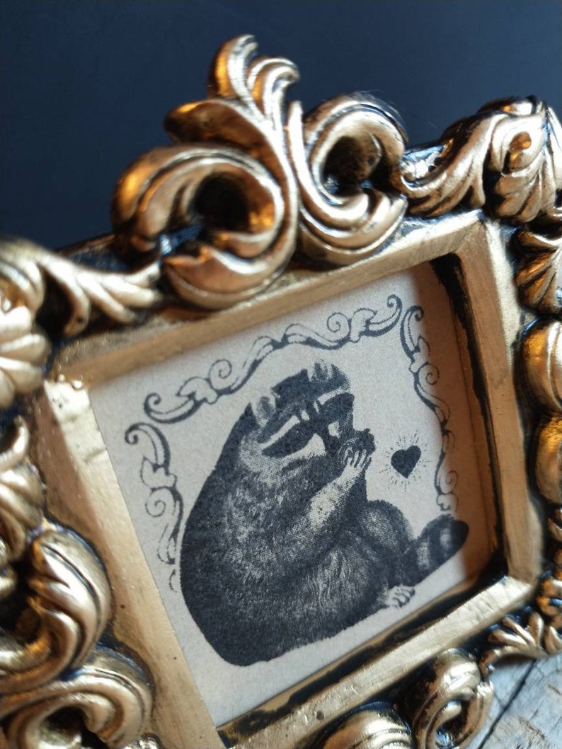 May include: An ornate picture frame in a gold colour with a black and white illustration of a raccoon holding a heart. The raccoon is sitting on a beige background with a black and white floral design.