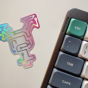 May include: A holographic sticker with a circuit board design in pink, blue, and blue colors. The sticker is on a white surface next to a gray mechanical keyboard with a green escape key.