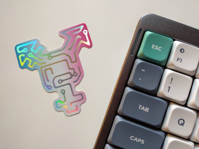 May include: A holographic sticker with a circuit board design in pink, blue, and blue colors. The sticker is on a white surface next to a gray mechanical keyboard with a green escape key.