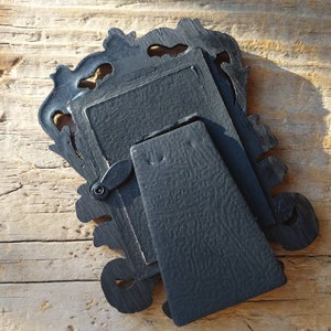 May include: Black ornate picture frame with a stand on the back. The frame is made of wood and has a detailed design.