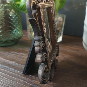 May include: A small, ornate, gold-colored picture frame with a black mat and a black and white photo inside. The frame has a decorative, ornate design with a black base.