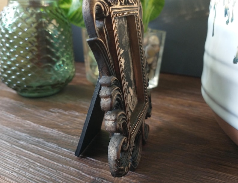 May include: A small, ornate, gold-colored picture frame with a black mat and a black and white photo inside. The frame has a decorative, ornate design with a black base.