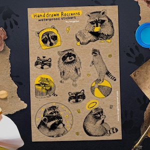 Puede incluir: Una hoja de pegatinas impermeables de mapaches dibujadas a mano. Las pegatinas presentan varias ilustraciones de mapaches con acentos amarillos, incluyendo mapaches con un sombrero de pan, equilibr&aacute;ndose sobre una pelota y comiendo. El texto dice "Handdrawn Raccoons" y "waterproof stickers".