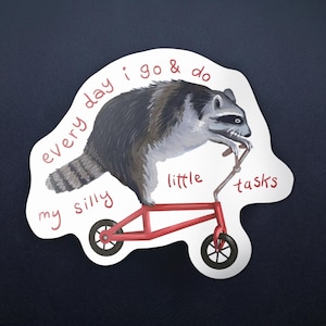 Puede incluir: Una pegatina con un mapache montado en un patinete rojo. El mapache es gris, negro y blanco. El texto de la pegatina dice "every day I go & do my silly little tasks."
