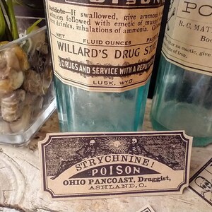 A scene showing antique reproduction label stickers from the Poison Bottle Labels sticker pack. The stickers are printed with blank ink on light brown kraft paper and are rectangular. Formaldehyde and arsenic stickers are shown on antique blue glass pharmacy bottles. Additional stickers such as strychnine are shown propped up on the bottle.