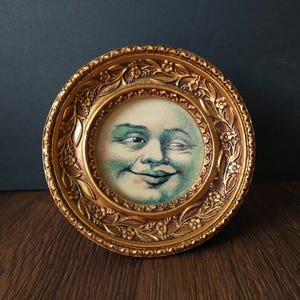 May include: A circular gold-toned frame with an ornate floral relief pattern surrounding a central circular print. The print features a stylized, pale-skinned face with green-tinted shading, depicting a moon-like figure with a subtle, knowing smile.