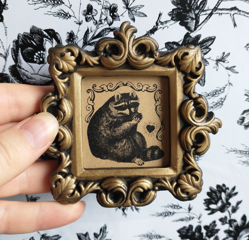 May include: An ornate picture frame in a gold colour with a black and white illustration of a raccoon holding a heart. The raccoon is sitting down and has a cute expression on its face.