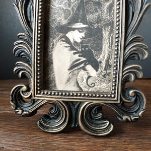 May include: A black and gold ornate picture frame with a black and white image of a witch holding a black cat. The witch is wearing a pointed hat and a long cloak.