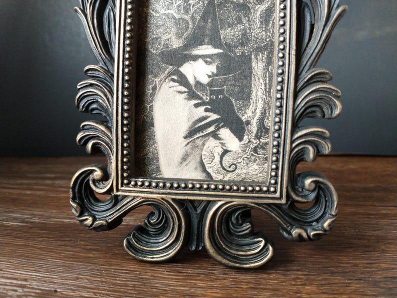 May include: A black and gold ornate picture frame with a black and white image of a witch holding a black cat. The witch is wearing a pointed hat and a long cloak.