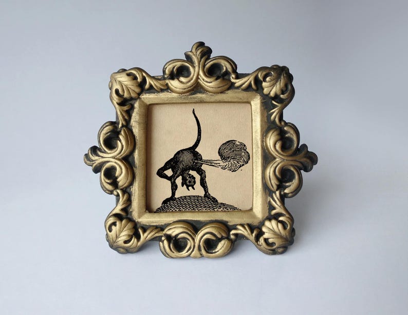 May include: A square picture frame with an ornate, gold-painted finish and dark recessed detailing. Inside the frame is a black and white illustration on a tan background depicting a small, horned creature bending over with a cloud of smoke.