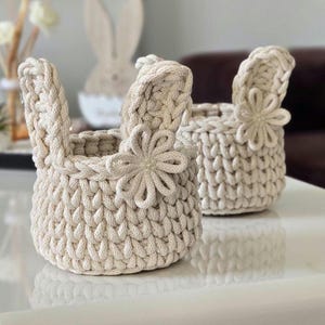 May include: Two beige crochet baskets shaped like bunnies. Each basket has a flower embellishment and bunny ears. The baskets are made of thick, textured yarn and sit on a reflective surface. The baskets are likely for Easter.