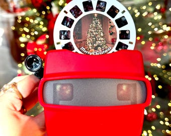 May include: A red View-Master toy with a white disc showing a Christmas tree scene and the words "Merry Christmas." The toy is held up against a background of Christmas lights.