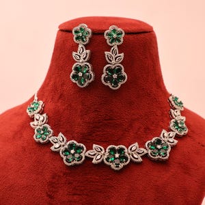 May include: A floral necklace and matching earrings set. The jewelry features green gemstones set in a silver-colored metal. The necklace and earrings are displayed on a red velvet bust against a light pink background.