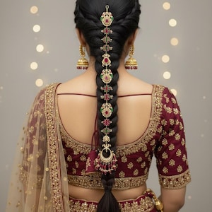 Puede incluir: Peinado trenzado de una mujer adornado con un accesorio decorativo para el cabello. El accesorio presenta adornos rojos, verdes y dorados. La mujer lleva un top bordado granate y pendientes dorados. El fondo tiene luces suaves y borrosas.