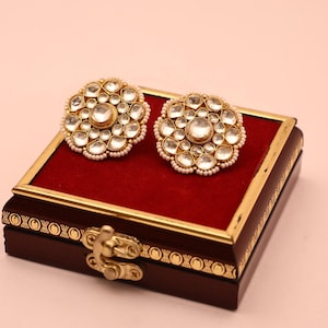 May include: A pair of gold-toned stud earrings with clear, faceted stones arranged in a floral pattern. The earrings are set on a red velvet-lined jewelry box with a gold trim and a decorative clasp. The box is dark brown.