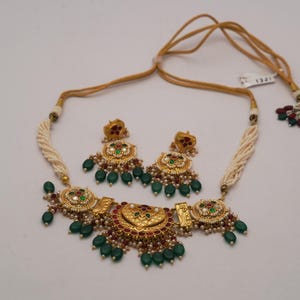 May include: A gold-coloured necklace and earring set with red, green, and white details. The necklace and earrings have intricate designs with green bead dangles and a pearl-like strand. The set is displayed against a white backdrop.
