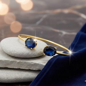 Puede incluir: Un brazalete dorado con dos grandes piedras preciosas ovaladas de color azul oscuro. El brazalete se exhibe sobre una pila de piedras grises sobre un fondo de tela de terciopelo azul oscuro. Las piedras preciosas son el foco de esta elegante joya.