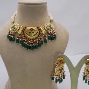 May include: A gold necklace and earring set. The necklace has a central pendant with red and green details, green teardrop beads and white pearls. The earrings match the necklace design.