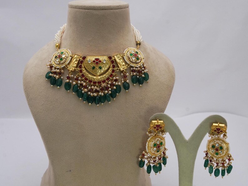 May include: A gold necklace and earring set. The necklace has a central pendant with red and green details, green teardrop beads and white pearls. The earrings match the necklace design.