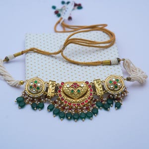 May include: An ornate gold necklace with red, green, and white embellishments. The necklace features a central heart-shaped design and is decorated with green bead dangles and white pearl strands. The necklace is displayed on a white and gold dotted surface.
