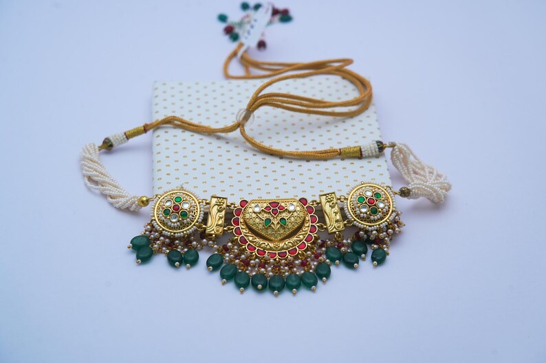 May include: An ornate gold necklace with red, green, and white embellishments. The necklace features a central heart-shaped design and is decorated with green bead dangles and white pearl strands. The necklace is displayed on a white and gold dotted surface.