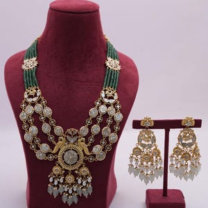 Puede incluir: Un conjunto de collar y pendientes de oro con un diseño de varias capas que presenta cuentas verdes, perlas blancas y detalles intrincados en oro. El colgante presenta un diseño circular con un motivo de pájaro.