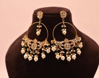 May include: Gold-toned hoop earrings with white and green accents and dangling faux pearls. The earrings feature a geometric design with a central white mosaic pattern. The earrings are displayed on a brown bust.