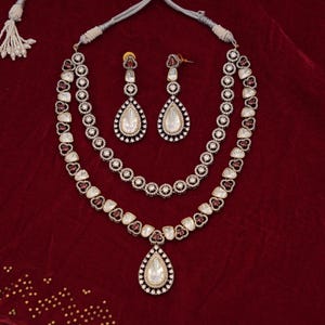 Puede incluir: Un collar de plata de dos niveles con un colgante en forma de lágrima. El collar está adornado con piedras blancas y rosas y tiene un cordón negro.