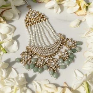 Puede incluir: Un tocado dorado adornado con hebras de perlas blancas y detalles de piedras preciosas verde claro. La joya presenta detalles intrincados y está rodeada de flores blancas, creando una estética elegante.