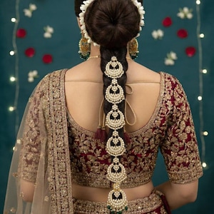 Peut inclure: Vue arrière d'une femme vêtue d'un haut et d'une jupe brodés de couleur bordeaux et or. Ses cheveux foncés sont coiffés en chignon avec une coiffe florale blanche. Un long accessoire capillaire orné de perles et d'or descend dans son dos.