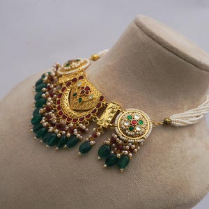 May include: An elaborate gold necklace with detailed patterns. It has emerald green beads, red embellishments, and tiny white pearls. The necklace is presented on a mannequin head, highlighting its design.