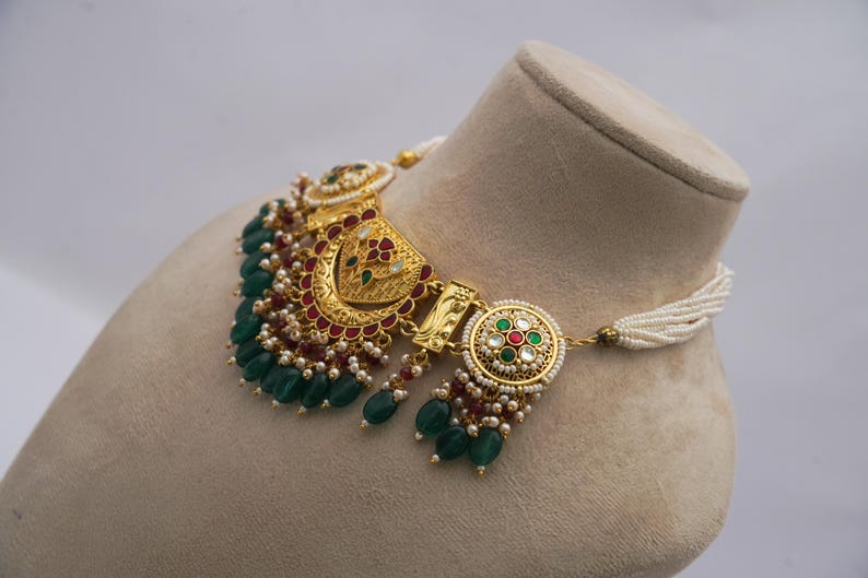 May include: An elaborate gold necklace with detailed patterns. It has emerald green beads, red embellishments, and tiny white pearls. The necklace is presented on a mannequin head, highlighting its design.