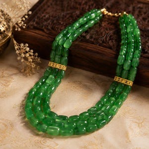 May include: A multi-strand necklace featuring vibrant green beads. The necklace has gold-colored accents and a gold clasp. The beads are a rich, emerald green color, and the necklace is displayed on a patterned surface.